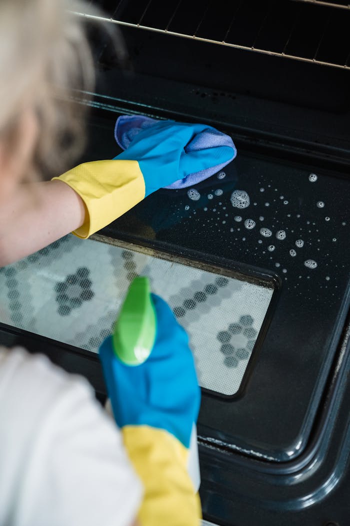 our-story A person wearing gloves cleans an oven door using a spray and cloth.
