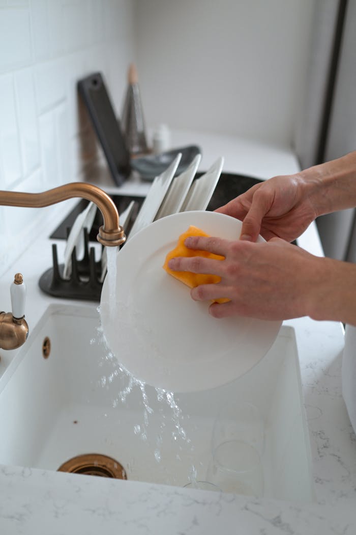 Hands washing a white dish in a modern kitchen sink with a yellow sponge.